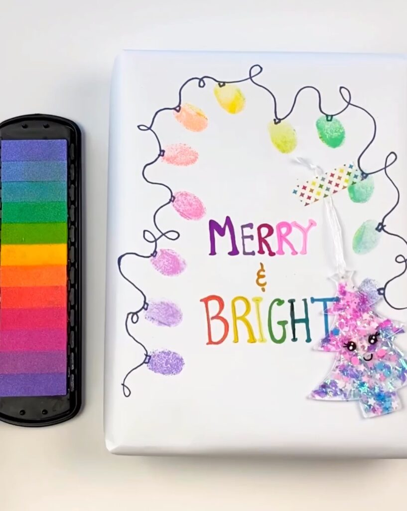 DIY Christmas Wrapping Paper - picture is of a wrapped gift that says "Merry & Bright" with stamped fingerprint lights and a rainbow stamp pad next to it