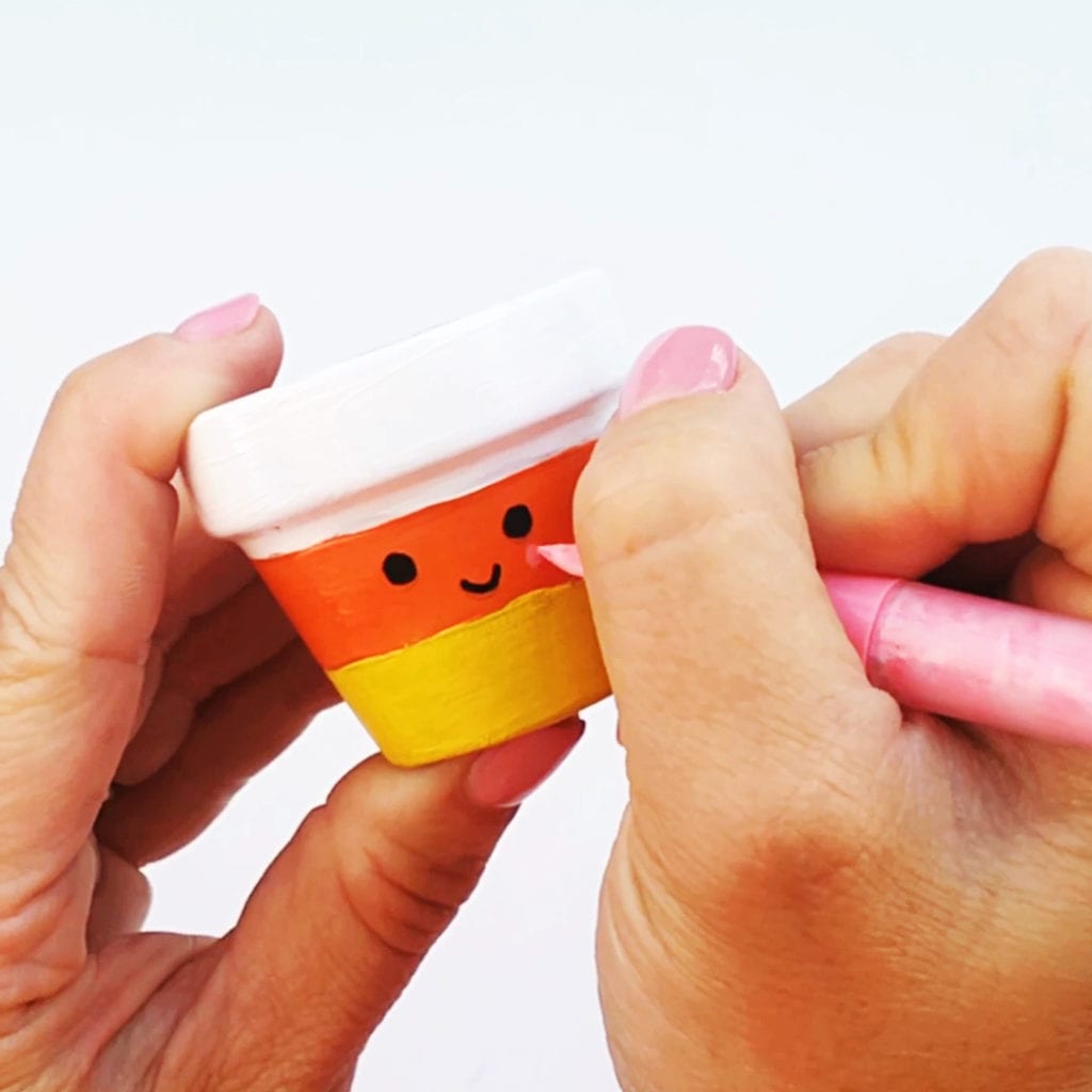 Adding cheeks with a paint marker to a candy corn design that's painted on a terracotta flower pot