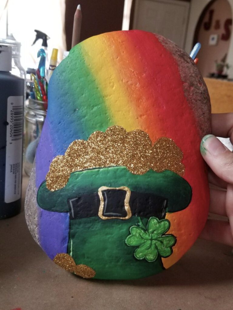 Upside down leprechaun hat with gold coins inside and a rainbow behind it painted onto a rock